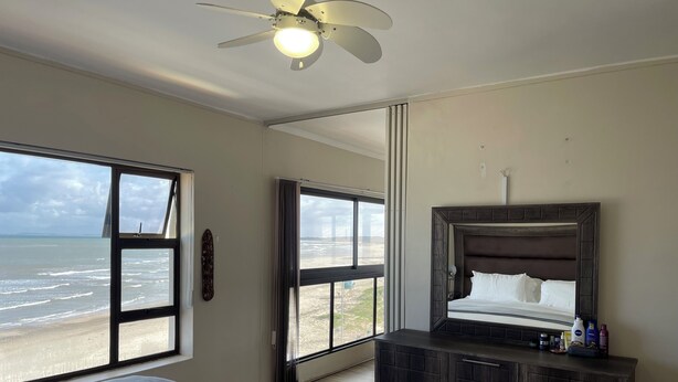 Ein Schlafzimmer mit Bett, Schminktisch und Deckenventilator. Große Fenster bieten Blick auf den Strand.