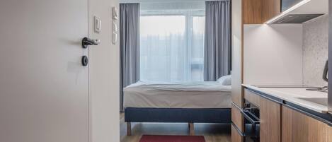 A modern hotel room with a bed, a small kitchenette, and a view of the city.
