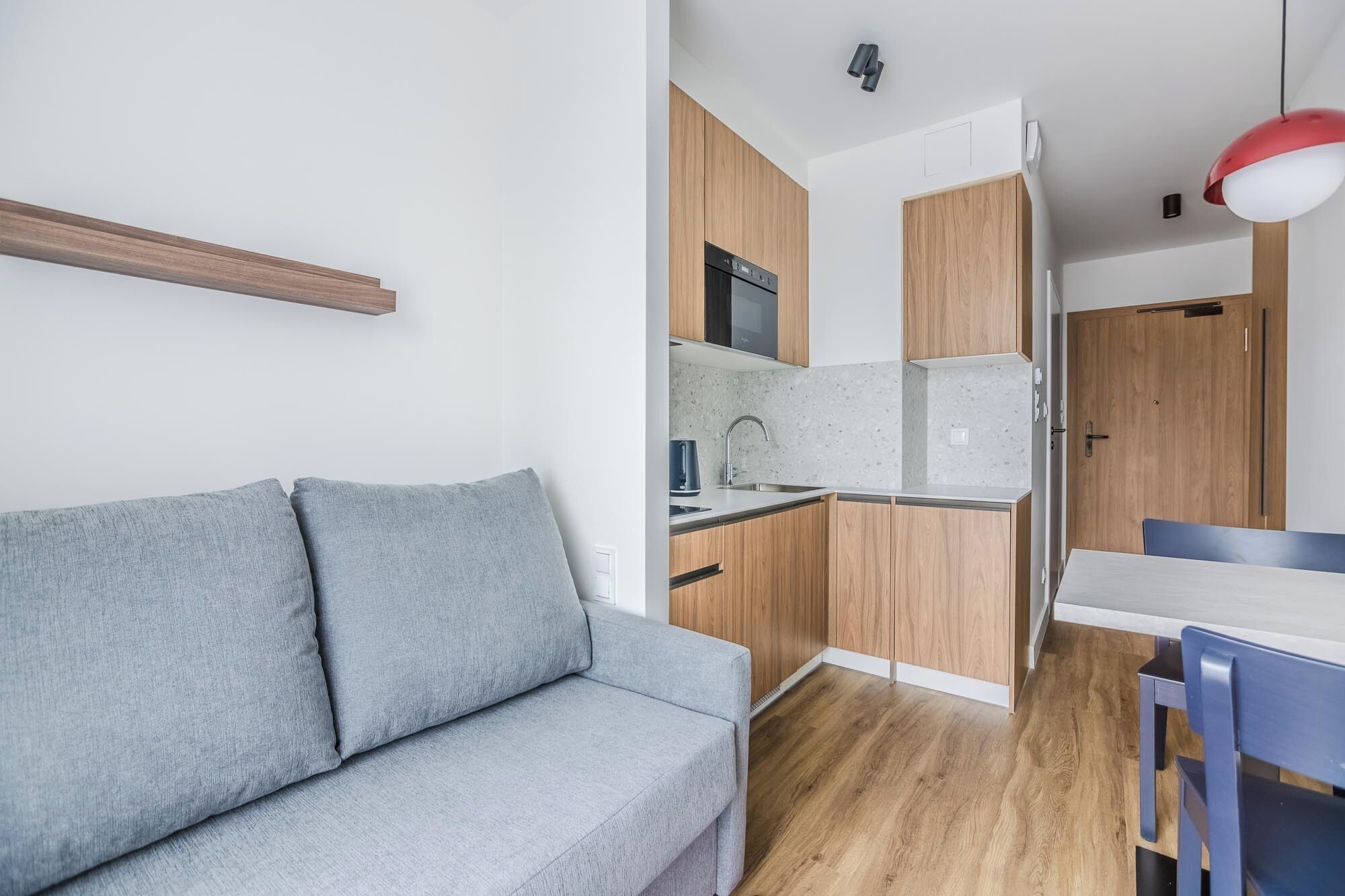 Un espacio habitable compacto con un sofá gris, una kitchenette con armarios de madera y una zona de comedor con una mesa y sillas.