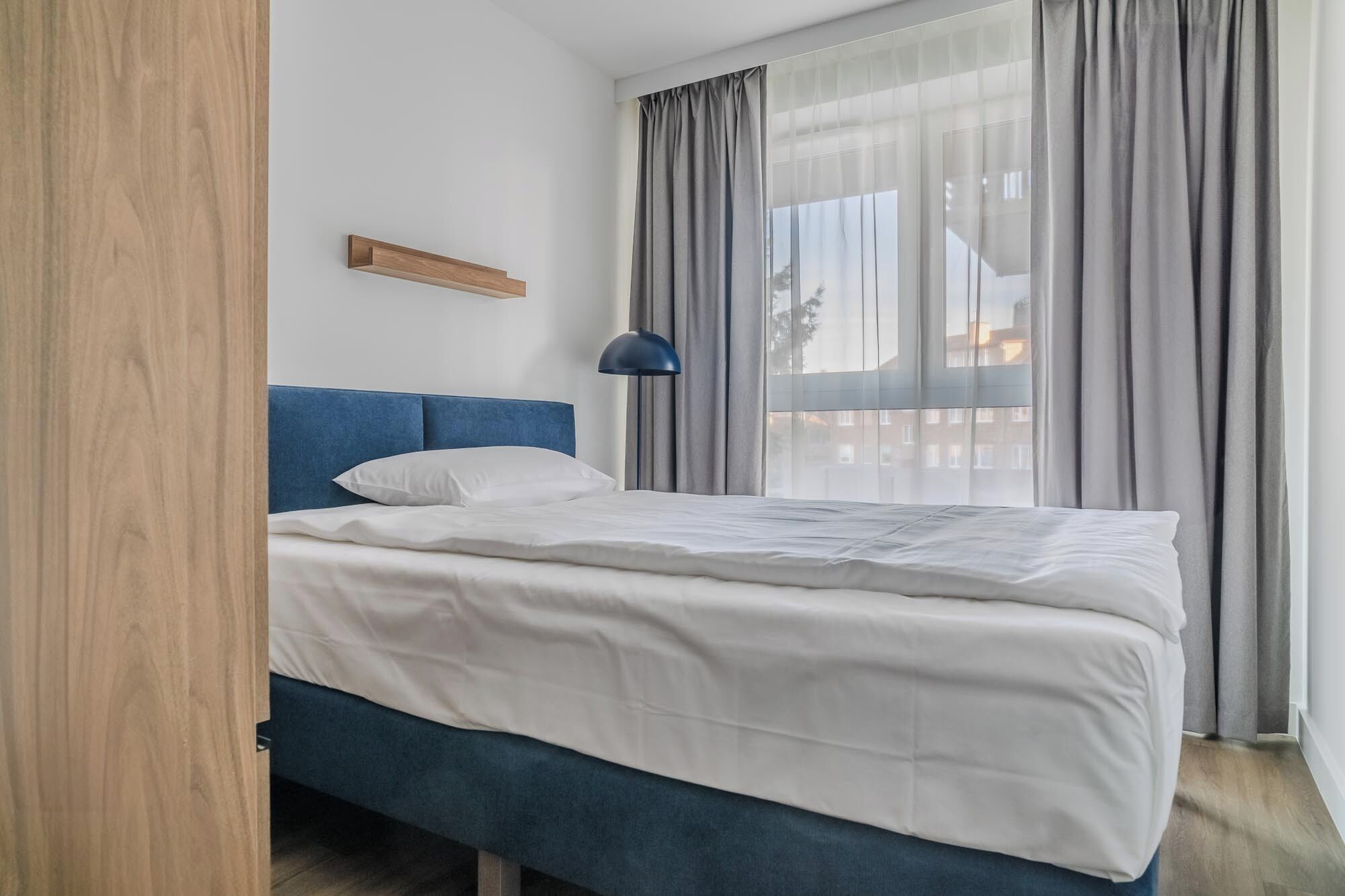 Un dormitorio con cama, cabecera azul, lámpara de noche y ventana con cortinas.