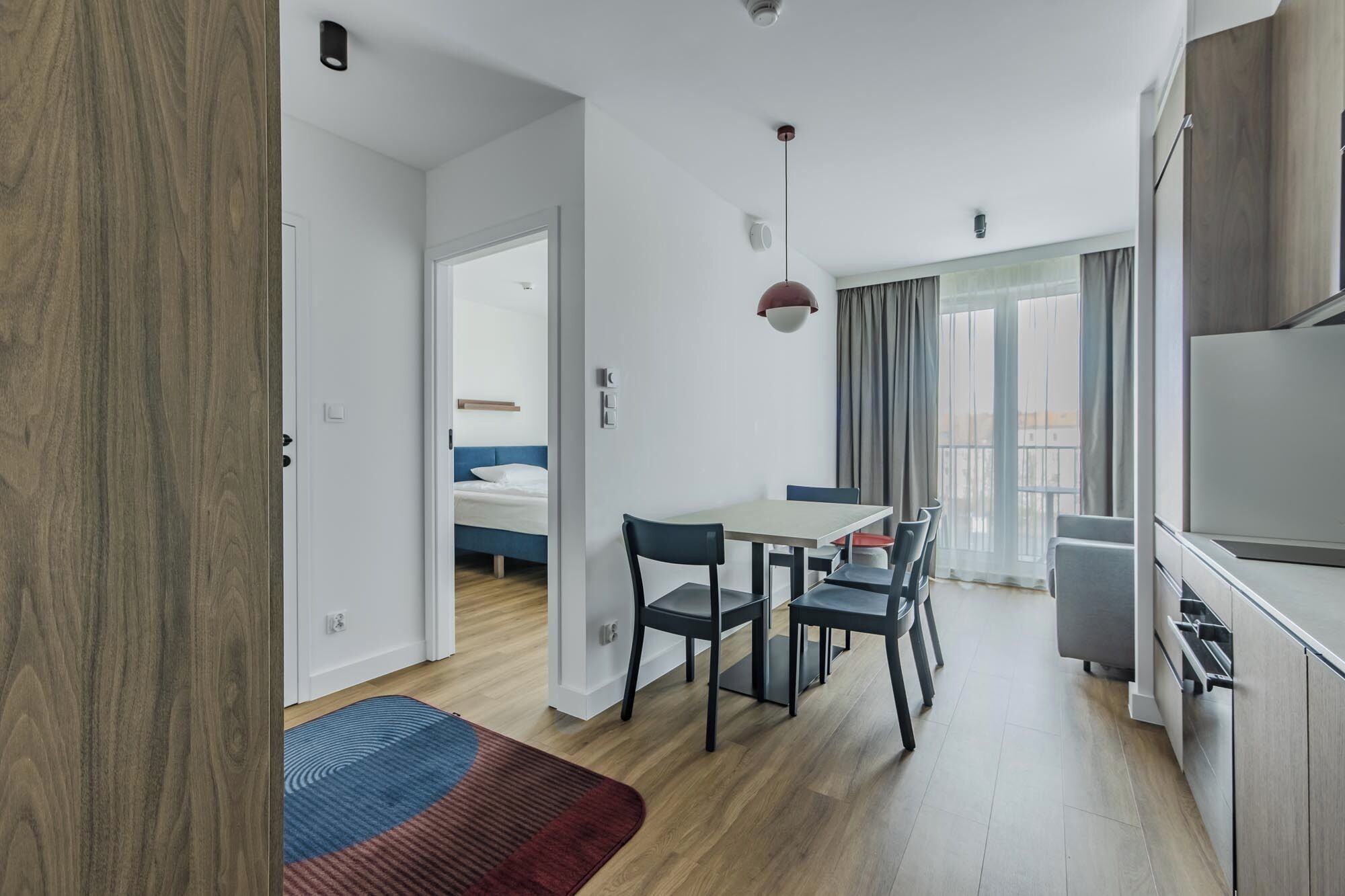 A modern hotel room with a wooden floor, a dining area with a table and chairs, a bed with a blue headboard, and a large window with curtains.