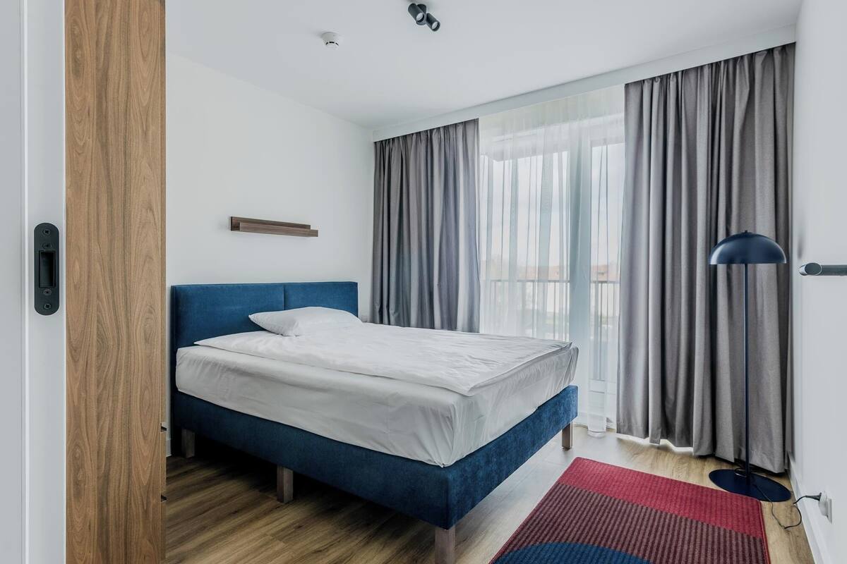 Een moderne slaapkamer met een blauw bed, witte beddengoed, een houten vloer en uitzicht op het landschap door grote ramen met grijze gordijnen.