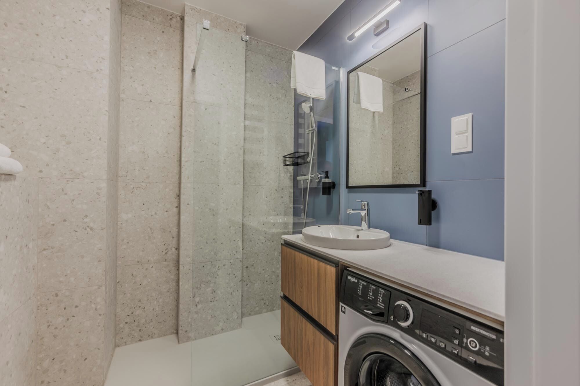 A modern bathroom with a glass shower enclosure, a sink, a mirror, and a washing machine.