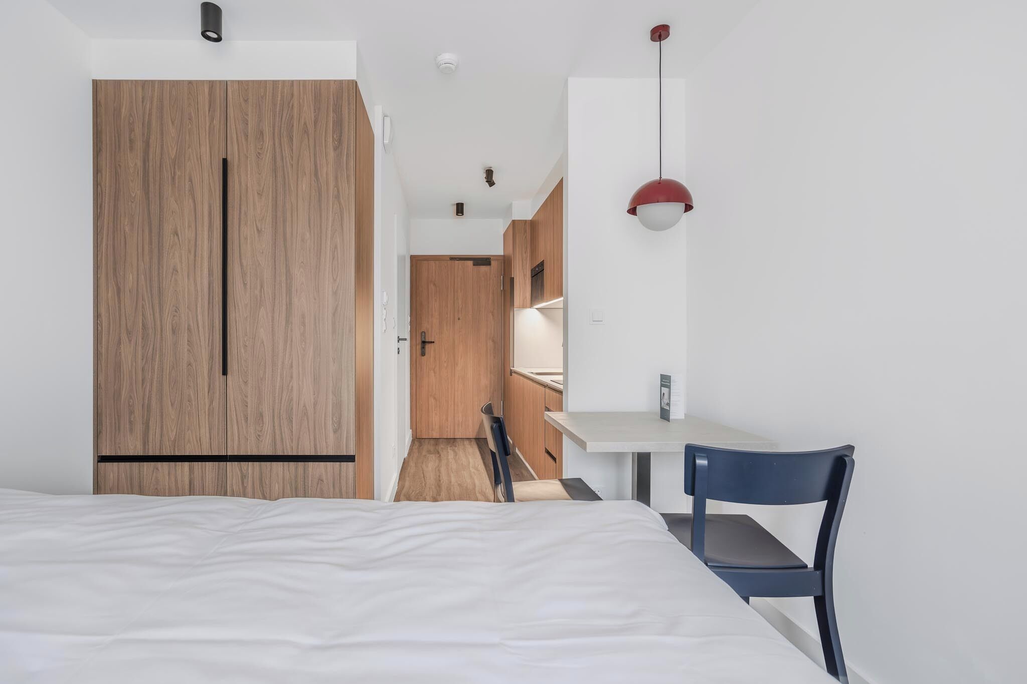A modern hotel room with a wooden wardrobe, a bed, a desk with a chair, and a pendant light.