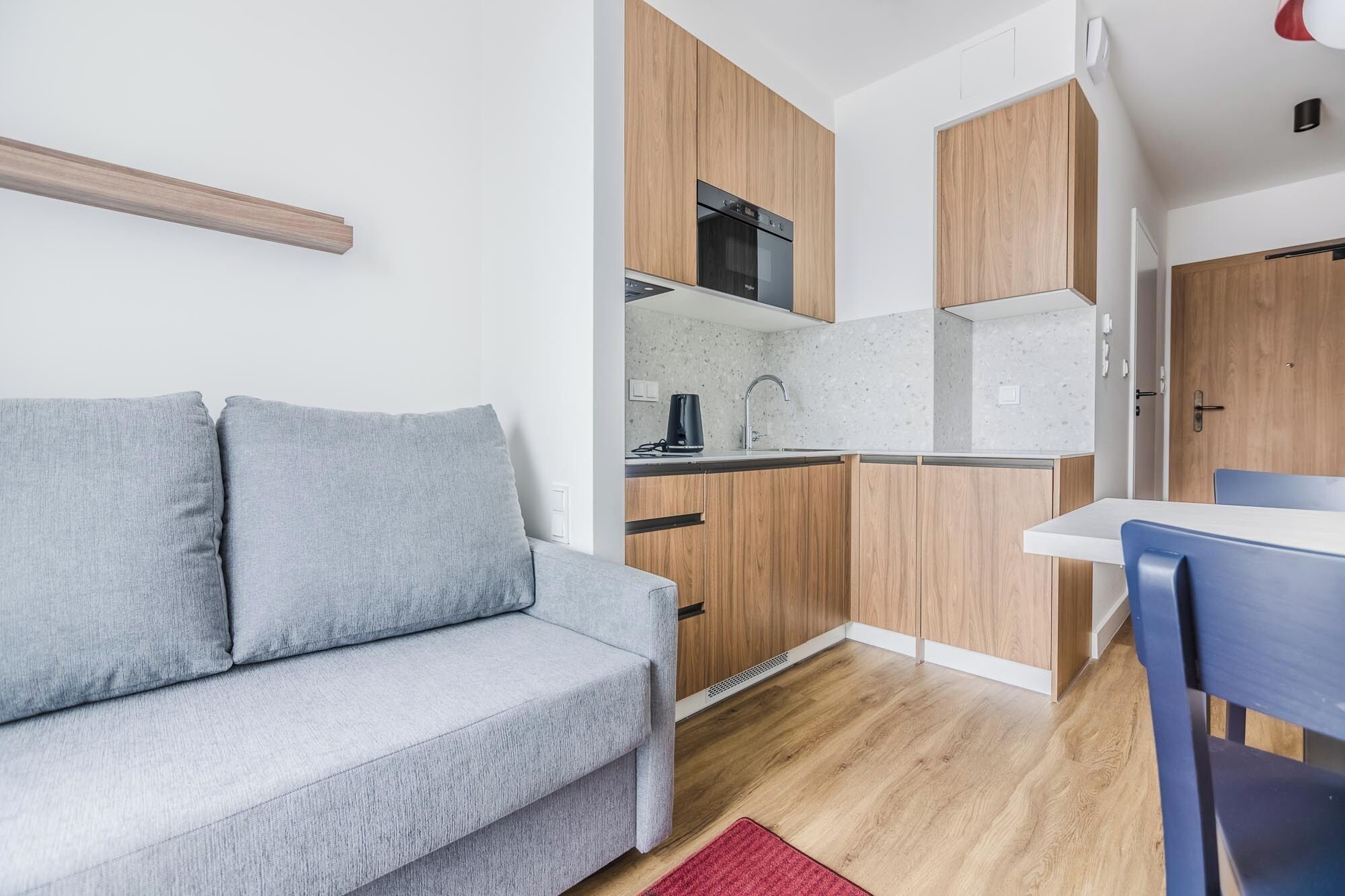 Un espacio habitable compacto con sofá gris, armarios de madera, una zona de cocina y una pequeña mesa de comedor.