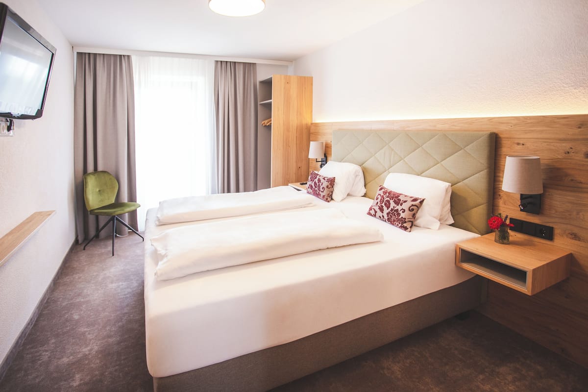 A hotel room with a large bed, two bedside tables, a green chair, a TV mounted on the wall, and a window with curtains.