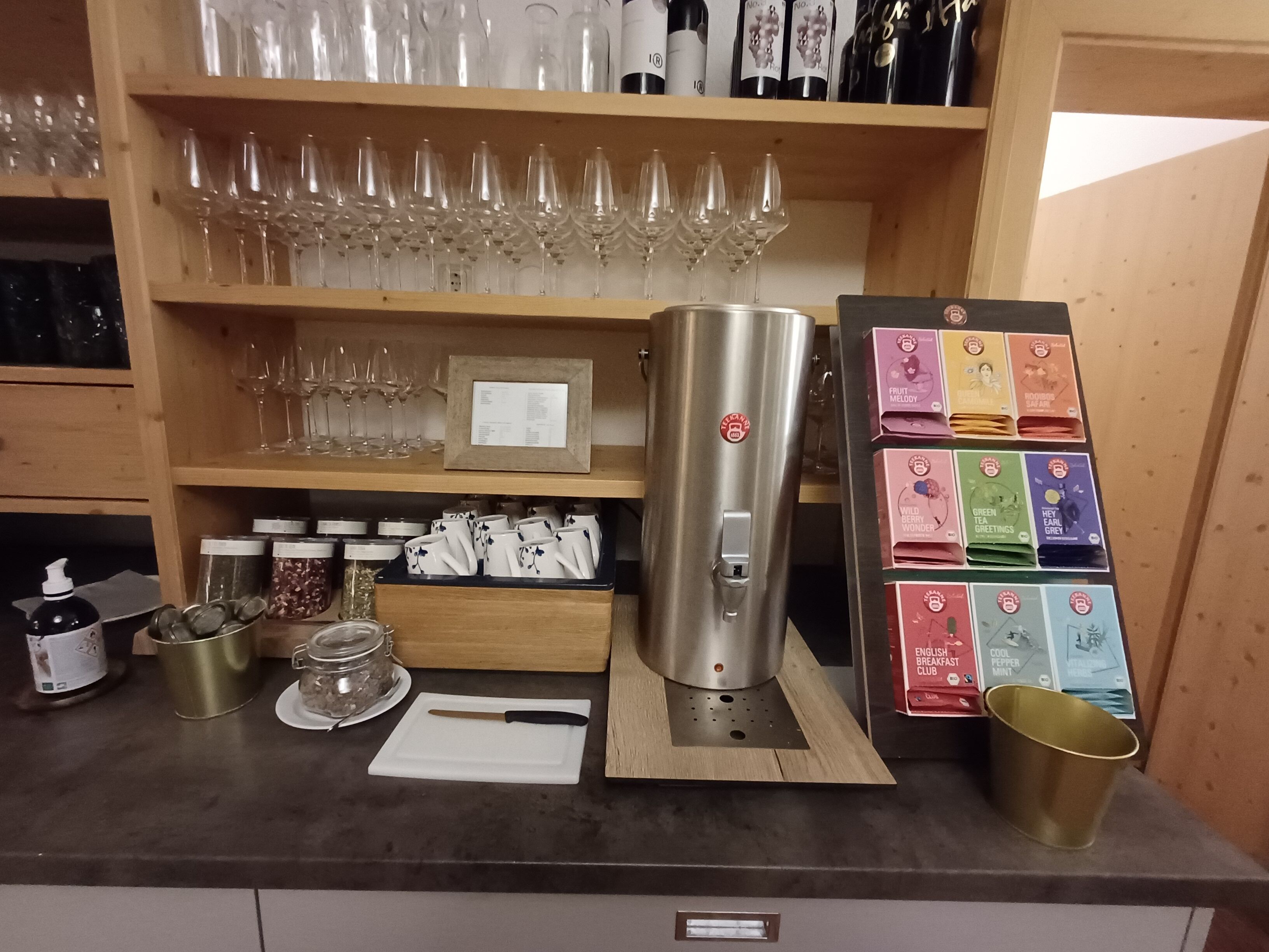 Een bar met wijnglazen, flessen, een koffieapparaat en theeboxen.