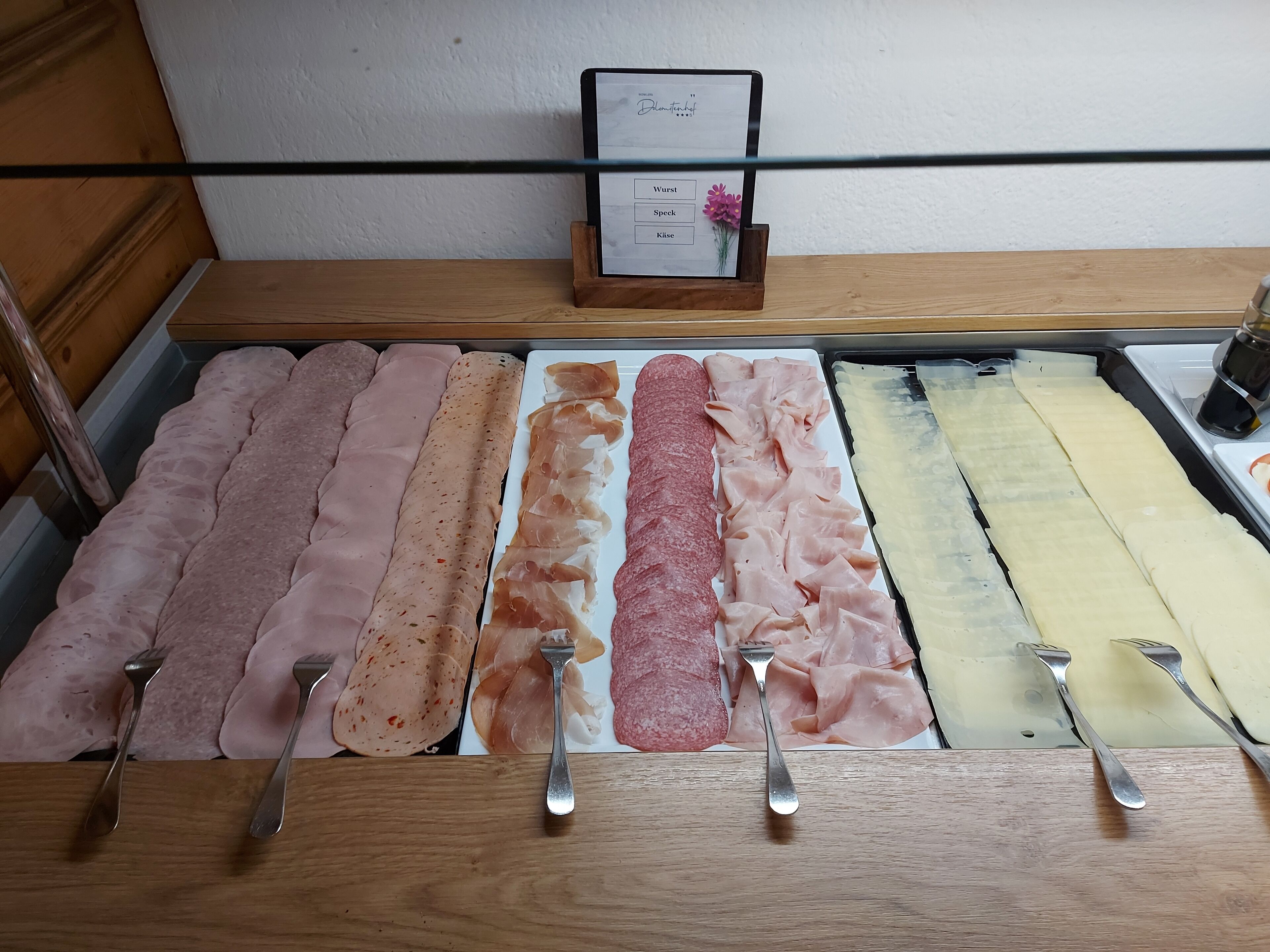 A deli counter with various types of deli meats and cheeses, including sliced ham, salami, and provolone, with forks placed in front.