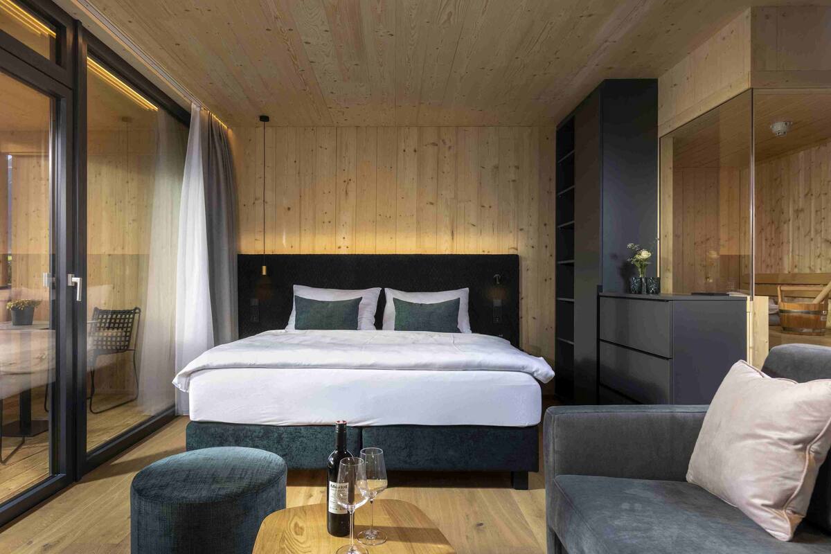A modern hotel room with a large bed, a wooden ceiling, a glass door leading to a balcony, and a small table with a bottle and glasses.