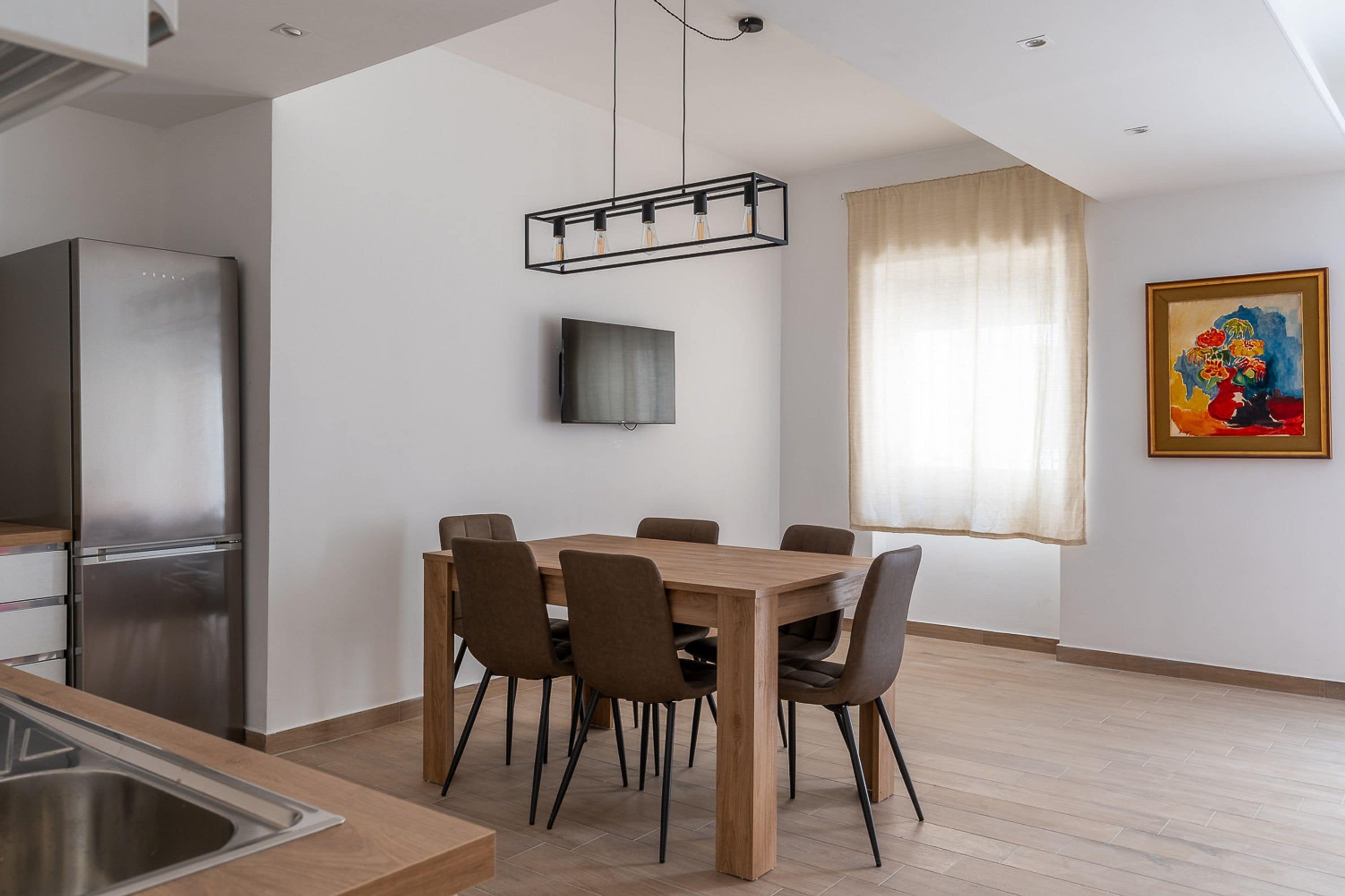 A modern kitchen with a stainless steel refrigerator, a wooden dining table with chairs, a hanging light fixture, a flat-screen TV, and a window with curtains.