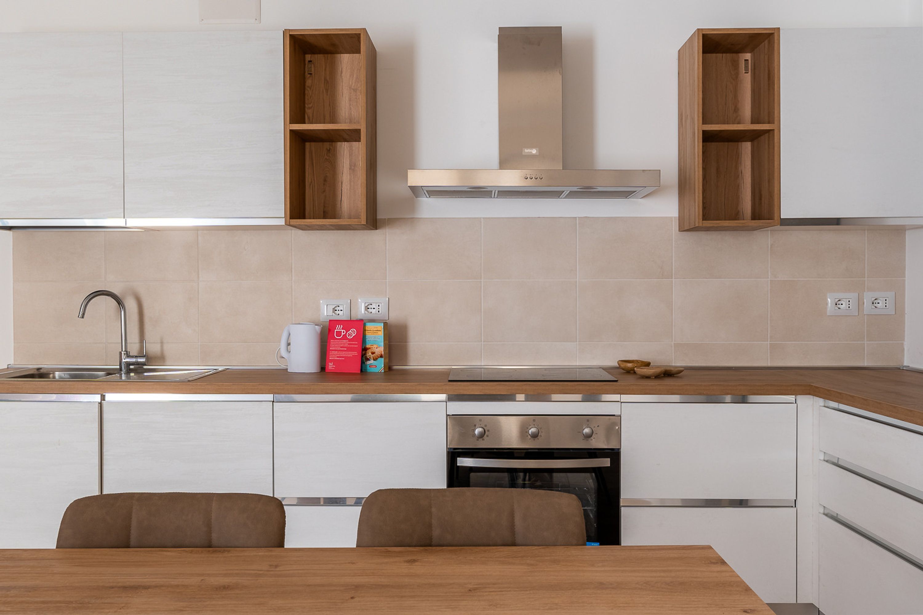 A modern kitchen with white cabinetry, a stainless steel range hood, and a built-in oven. There are two wooden cabinets above the sink and a dining table with chairs.