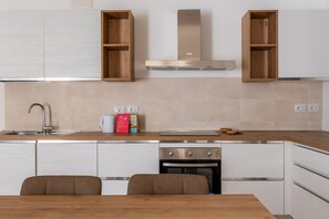 A modern kitchen with white cabinetry, a stainless steel range hood, and a built-in oven. There are two wooden cabinets above the sink and a dining table with chairs.