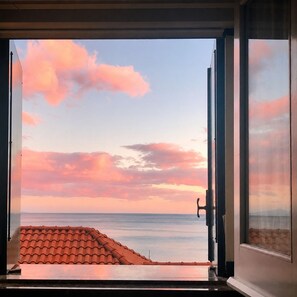 Une porte ouverte donnant sur un coucher de soleil sur la mer, un toit en tuiles et une rambarde en métal.