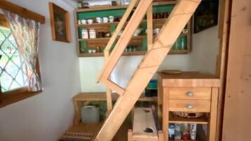 Ein Zimmer mit einer Holztreppe, Regalen mit verschiedenen Gegenständen, einem kleinen Tisch und einem Fenster mit gemusterten Vorhängen.