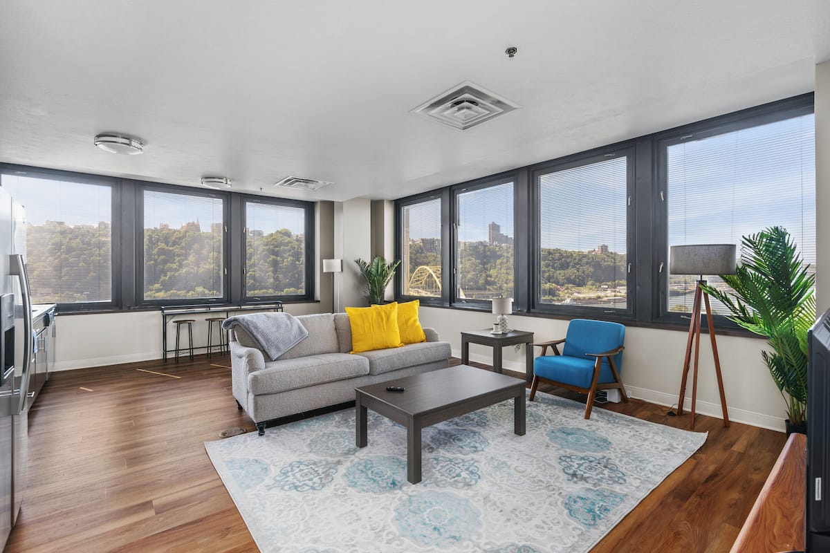 Ein modernes Wohnzimmer mit Sofa, Couchtisch und Blick auf eine Stadtlandschaft durch große Fenster.