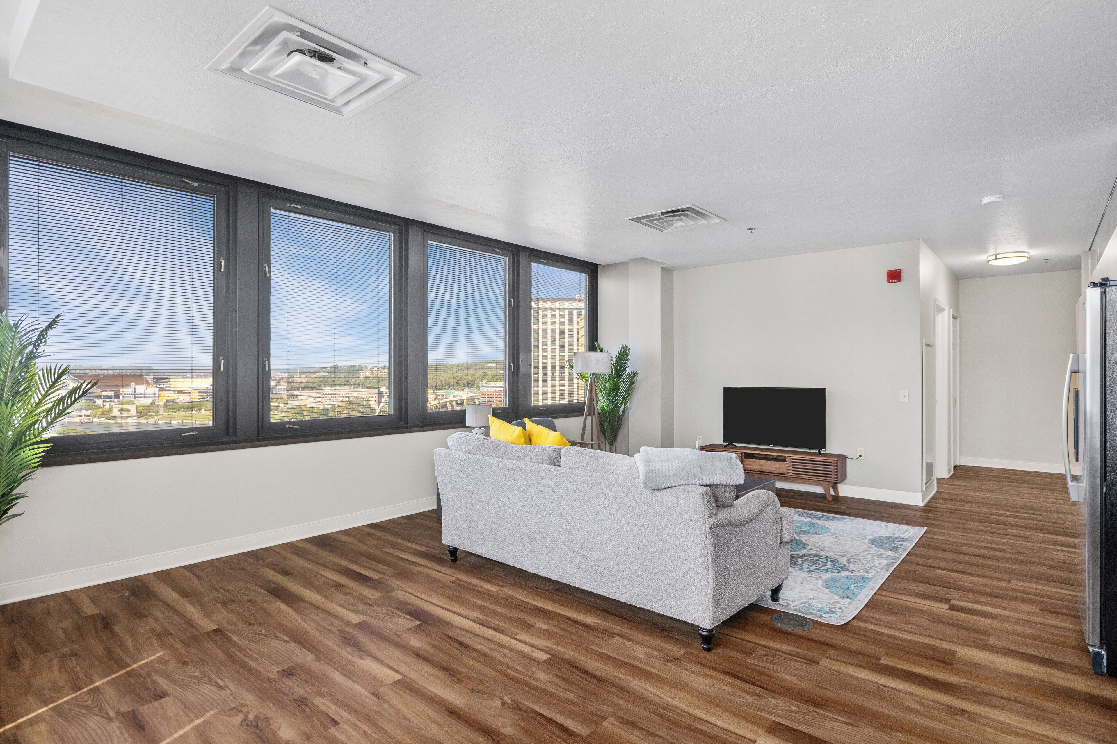 A modern living room with a grey sofa, a flat-screen TV, and a large window offering a city view.