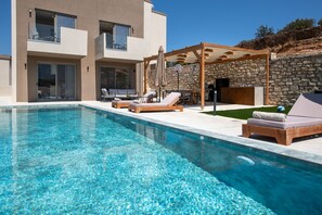 A modern pool area with lounge chairs and a pergola, surrounded by a stone wall and a two-story building. - Calmo Luxury Villa II, with Pool Tranquil Elegance (9PXG+6WX Geropotamos, Greece)