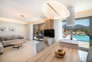 A modern living room with a sofa, coffee table, and dining table. There is a TV, a large window with a pool view, and a ceiling light fixture. - Calmo Luxury Villa II, with Pool Tranquil Elegance (9PXG+6WX Geropotamos, Greece)