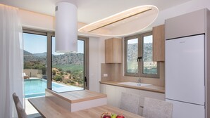 Une cuisine moderne avec un plan de travail en bois, un four encastré et un réfrigérateur. On y trouve une table à manger avec un panier de fruits, et la vue donne sur une piscine et des collines en arrière-plan.