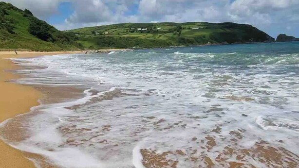 A beach with waves lapping onto the shore, a clear sky, and green hills in the background.