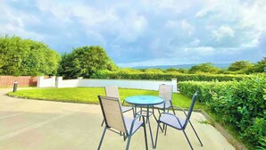 An outdoor patio area with a round table and four chairs, surrounded by well-maintained hedges and a view of distant hills.