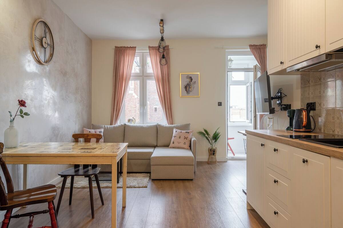Un salon confortable avec un canapé, une table à manger et des chaises. Il y a un coin cuisine avec des placards et une machine à café. Une horloge est accrochée au mur et une plante est posée sur le sol.