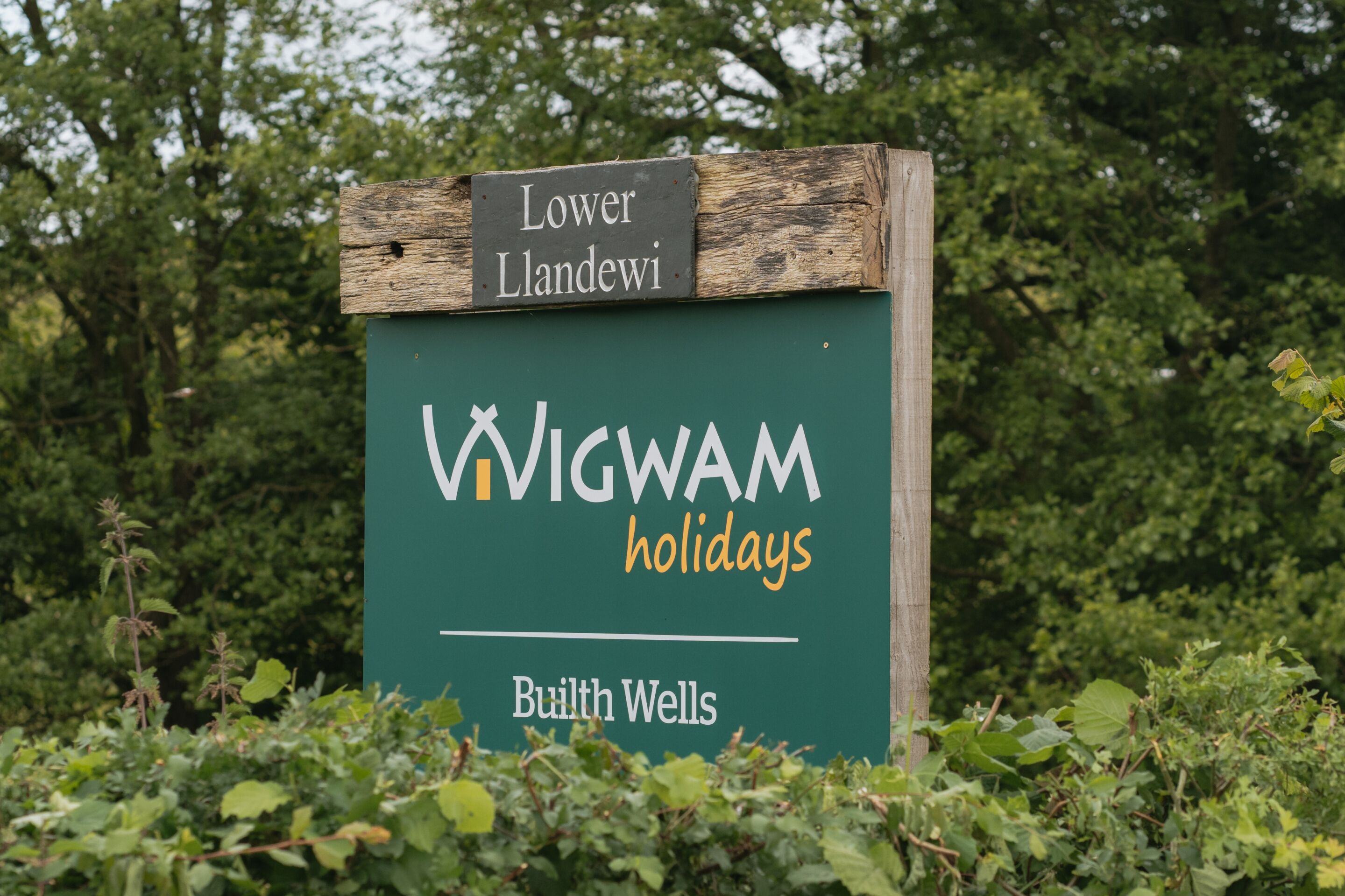 Een bord voor Wigwam Holidays met een rustieke houten bovenkant en een groene achtergrond.