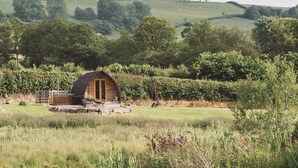 A rustic cabin with a curved roof, surrounded by lush greenery and rolling hills. - Cosy En-suite Glamping Cabin with Hot Tub (Erwood)