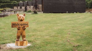 A wooden bear sculpture holding a sign that says "Welcome" in a grassy area with a wooden building in the background. - Cosy En-suite Glamping Cabin with Hot Tub (Erwood)