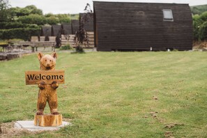 Una escultura de madera de un oso sosteniendo un letrero que dice 'Bienvenido' en un área cubierta de césped, con un edificio de madera al fondo.