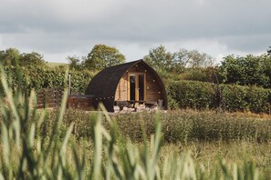 A wooden cabin with a sloped roof, surrounded by greenery and a fence. - Cosy En-suite Glamping Cabin with Hot Tub (Erwood)