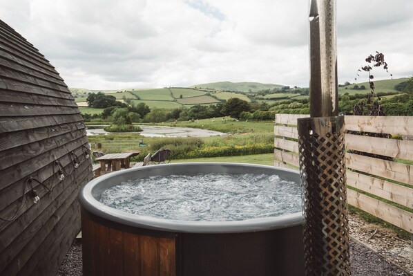 A hot tub in a rustic outdoor setting with a wooden cabin and a view of rolling hills in the background.