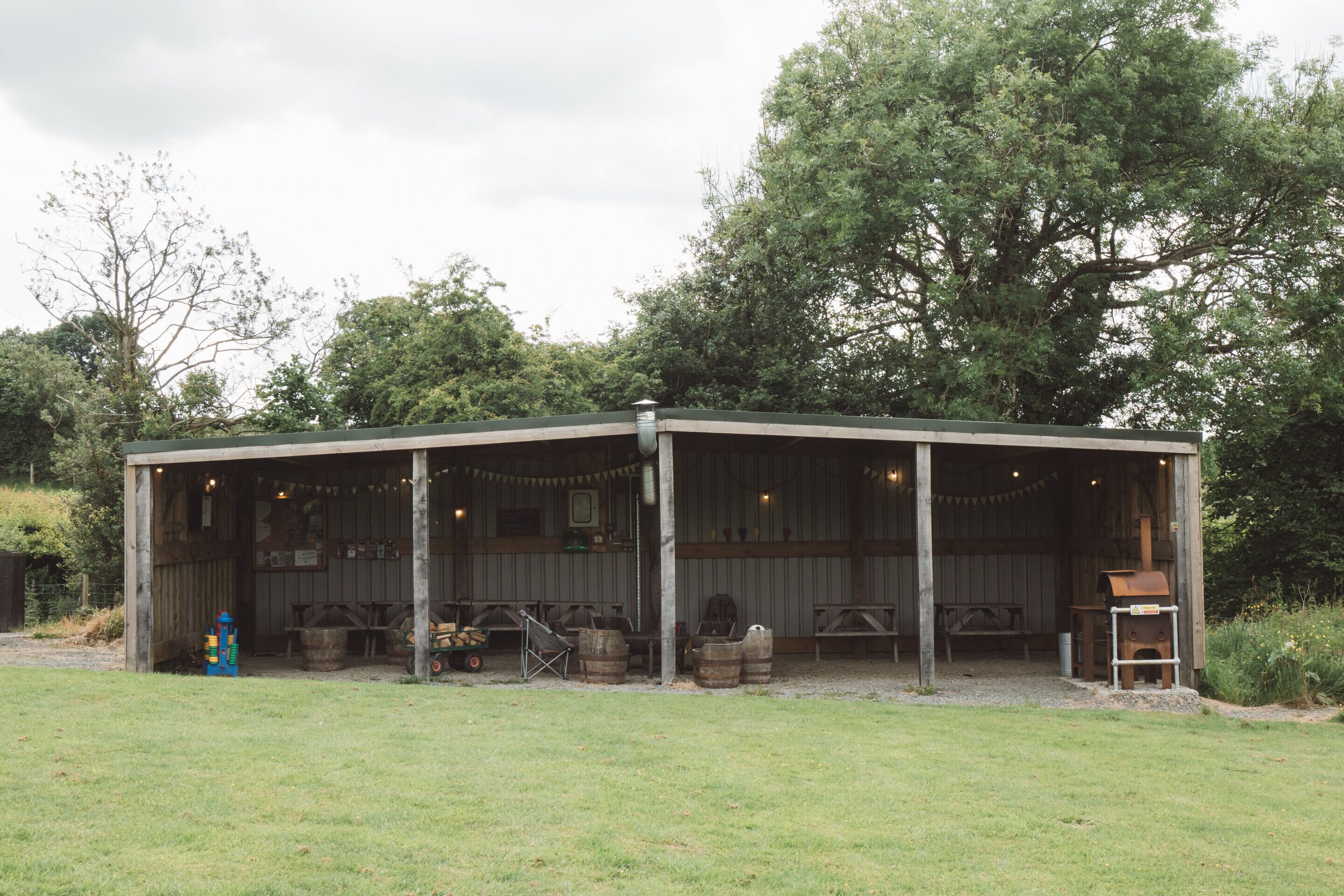 A rustic outdoor pavilion with a covered seating area, wooden barrels, and a grill.