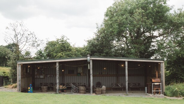 A rustic outdoor pavilion with a covered seating area, wooden barrels, and a grill. - Cosy En-suite Glamping Cabin with Hot Tub (Erwood)