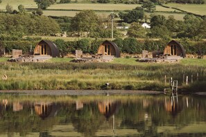 A row of wooden cabins with outdoor seating, a pond in the foreground, and a rural landscape in the background. - Cosy En-suite Glamping Cabin with Hot Tub (Erwood)