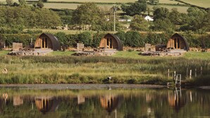 A row of wooden cabins with outdoor seating, a pond in the foreground, and a hilly landscape in the background. - Cosy En-suite Glamping Cabin Near Hay-on-Wye. (Erwood)