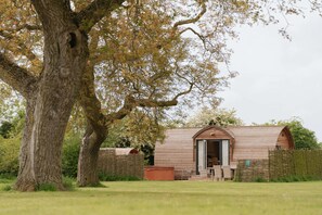 A rustic wooden cabin with a sloped roof, a wooden door, and a small porch area with a chair and table. - Cosy Rural Cabin With Hot Tub (Spalding)