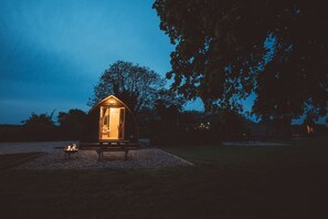 Una pequeña estructura iluminada con un banco en frente, rodeada de grava y árboles.