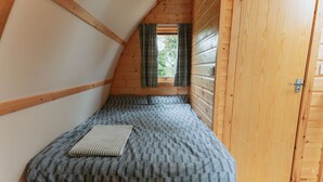 Una habitación pequeña de madera con techo inclinado, una cama individual con colcha estampada, una ventana con cortinas y una puerta de madera.
