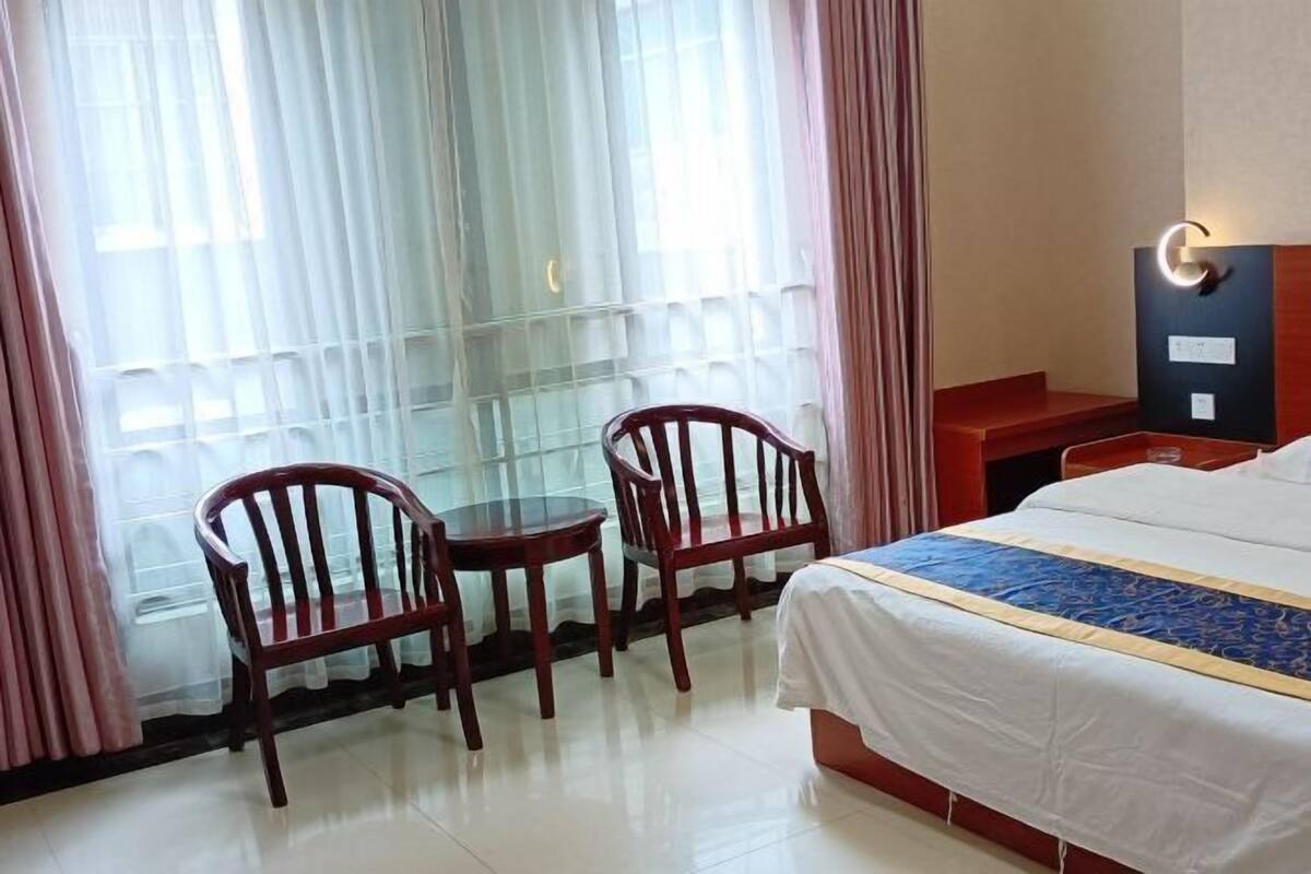 A hotel room with a bed, two wooden chairs, a small table, a nightstand, and a window with sheer curtains.