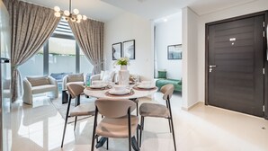 Uma sala de jantar moderna com uma mesa redonda posta para quatro pessoas, rodeada por cadeiras bege. Há um candelabro sobre a mesa, e o espaço possui uma janela grande com cortinas.
