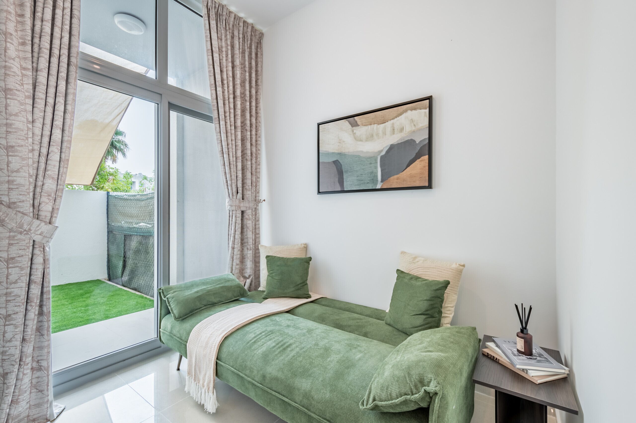 A modern bedroom with a green sofa bed, a small bedside table with a book and a candle holder, and a large window with curtains.