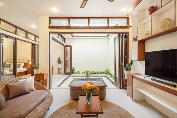 A modern living room with a sofa, a coffee table with a vase of flowers, a TV, and a view of a garden through a glass door. - The Seven Residence (Kerobokan)