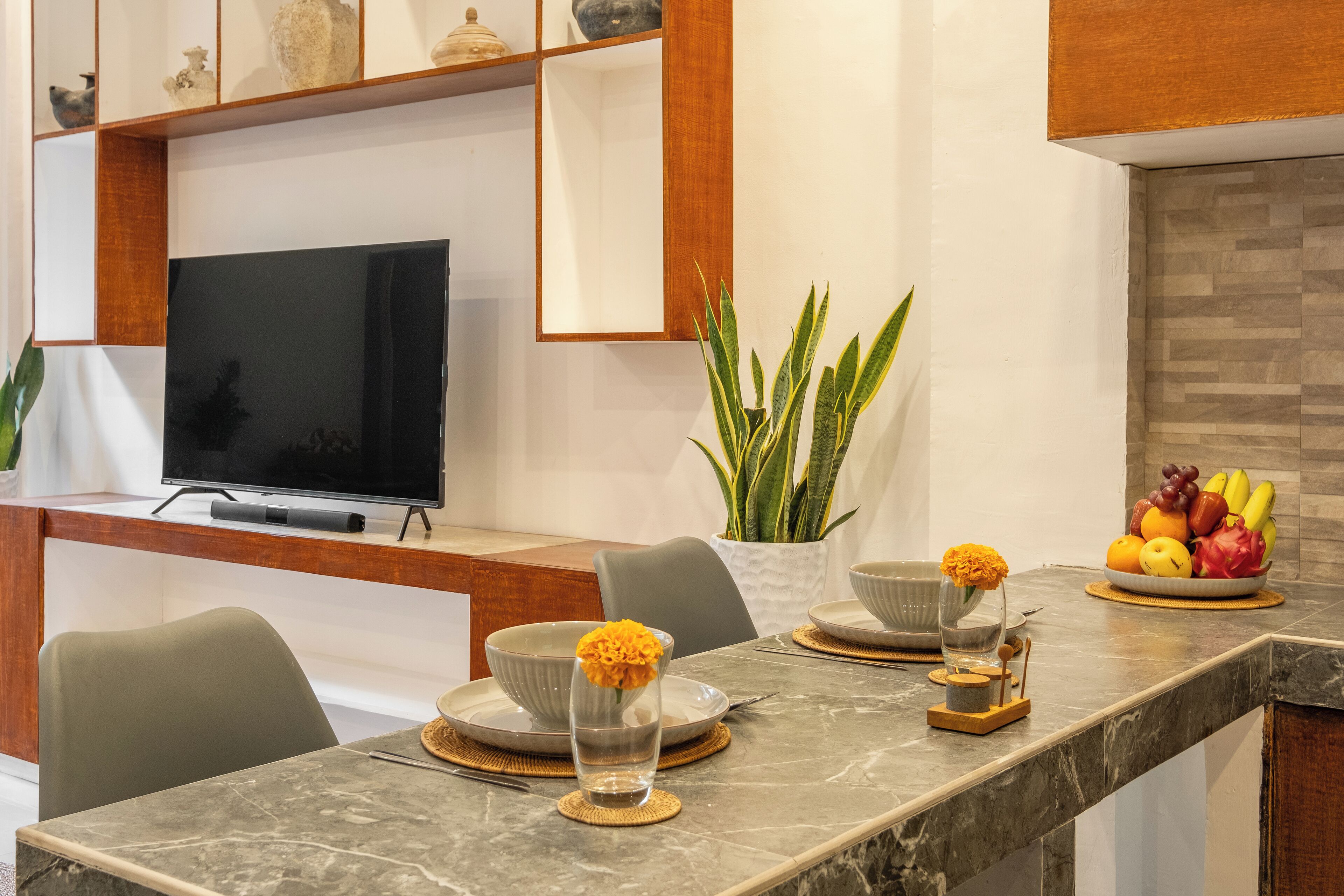 A modern kitchen with a marble countertop, a flat-screen TV, and a fruit bowl.
