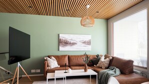Een moderne woonkamer met een bruine leren bank, een witte salontafel, een flatscreen televisie op een driepootstandaard en een groot raam met zonwering.