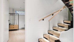 Een moderne trap met houten treden en glazen leuningen, leidend naar een kamer met een witte wand en een houten dressoir.