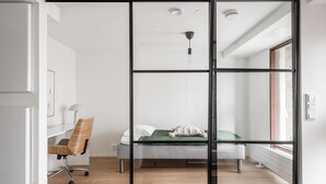Een moderne slaapkamer met een glazen scheidingswand, een bed, een bureau met stoel, en een raam met jaloezieën.