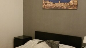 A neatly made bed with a patterned bedspread and pillows, a small bedside table with a lamp, and a framed picture of a cityscape on the wall. - Affittacamere Guerrero Jr (Trieste)