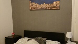 A bedroom with a bed, a nightstand, a lamp, a picture of a cityscape, and a vase with flowers. - Affittacamere Guerrero Jr (Trieste)
