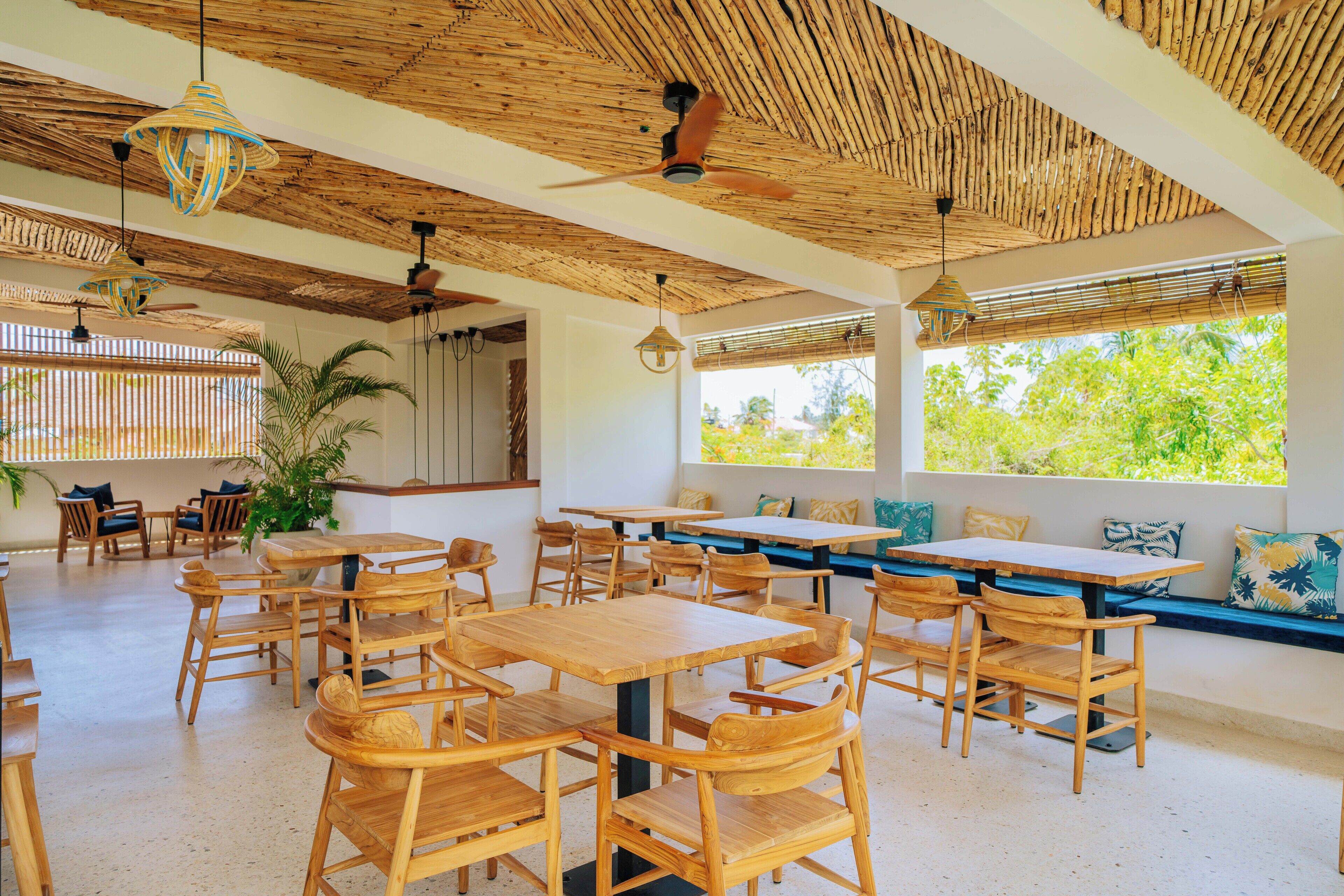 Un espace intérieur spacieux, doté de tables et de chaises en bois, d’un ventilateur de plafond et de grandes fenêtres offrant une vue sur la verdure environnante.